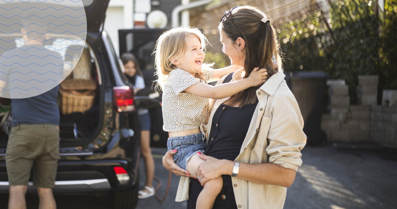 Frau hält ein lachendes Kind auf dem Arm, im Hintergrund packt ihr Mann das Auto. 