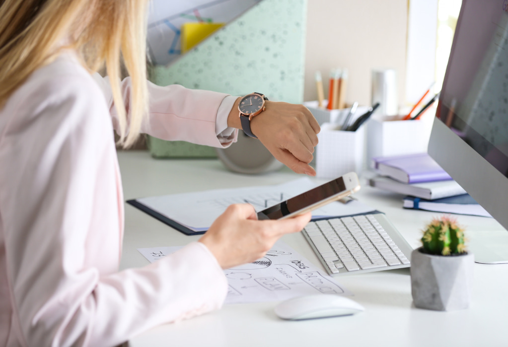 Eine Frau sitzt an einem Schreibtisch. Sie hat ihr Handy in der Hand und guckt auf ihre Armbanduhr. 