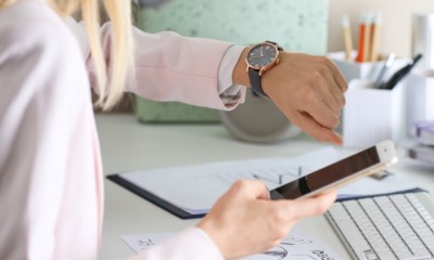 Eine Frau sitzt an einem Schreibtisch. Sie hat ihr Handy in der Hand und guckt auf ihre Armbanduhr. 