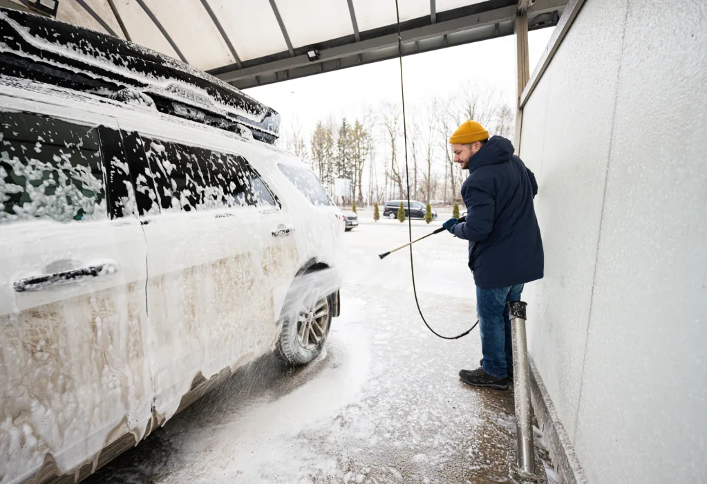 Ein Mann wäscht im Winter ein verschneites Auto mit einem Hochdruckreiniger in einer Waschanlage.