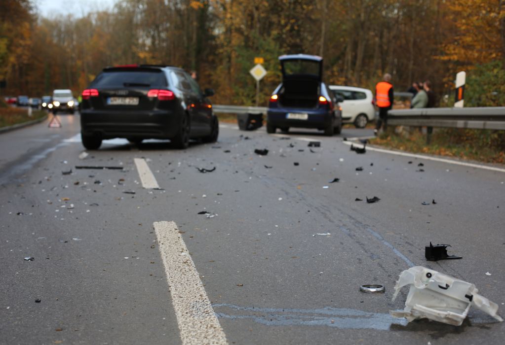 Ein Verkehrsunfall auf einer Landstraße mit zwei beteiligten Autos. Auf der Straße liegen Trümmerteile. Im Hintergrund stehen mehrere Personen mit Warnwesten.
