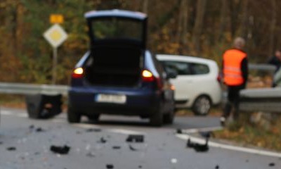 Ein Verkehrsunfall auf einer Landstraße mit zwei beteiligten Autos. Auf der Straße liegen Trümmerteile. Im Hintergrund stehen mehrere Personen mit Warnwesten.