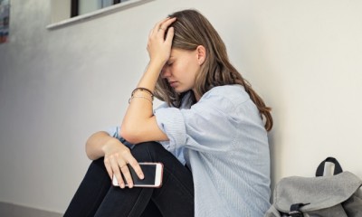 Ein junges Mädchen sitzt auf dem Fußboden mit ihrem Handy in der Hand und stützt den Kopf in die Hände.