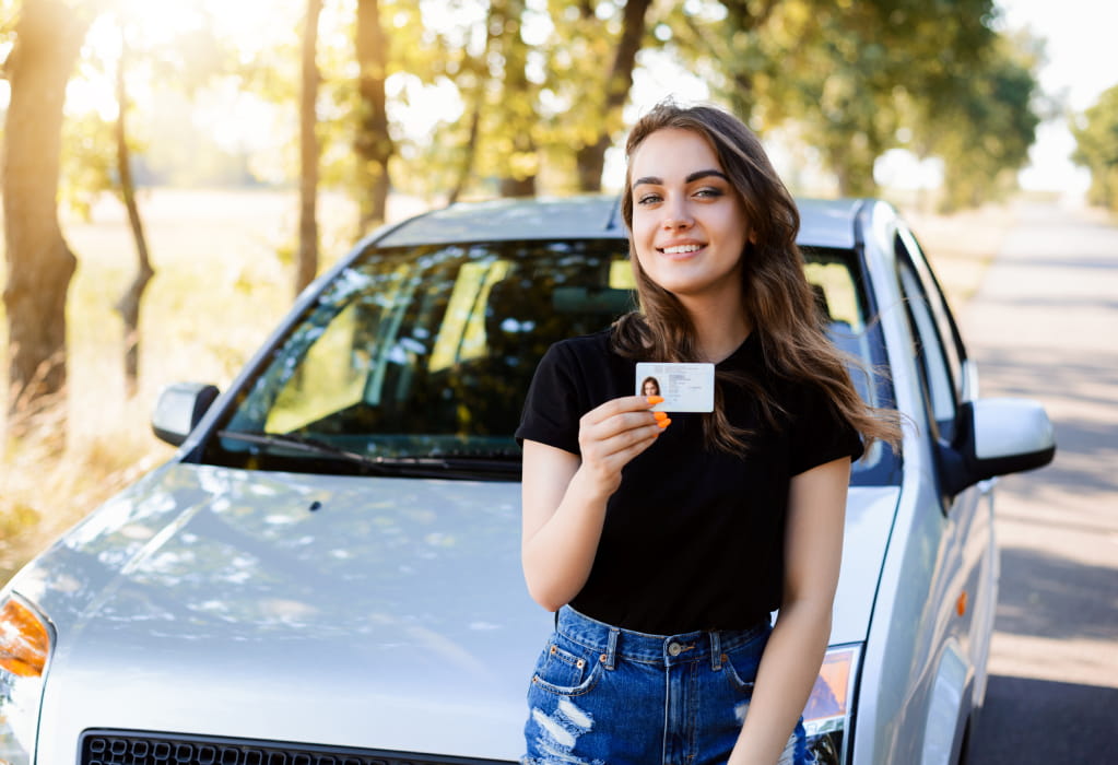 Eine junge Frau steht vor einem Auto und hält ihren Führerschein ins Bild.