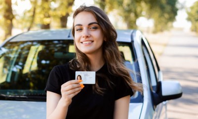 Eine junge Frau steht vor einem Auto und hält ihren Führerschein ins Bild.