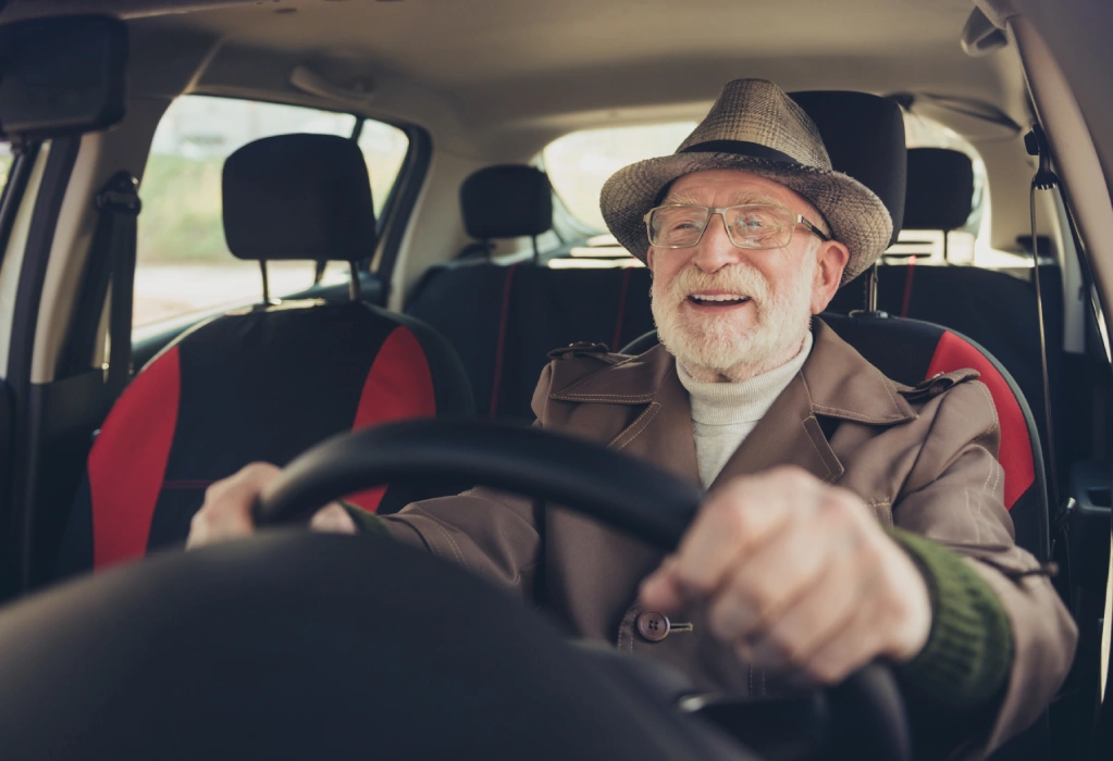 Ein lächelnder älterer Mann mit Brille und Hut sitzt am Steuer eines Autos und fährt.