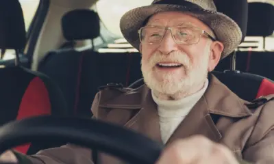 Ein lächelnder älterer Mann mit Brille und Hut sitzt am Steuer eines Autos und fährt.