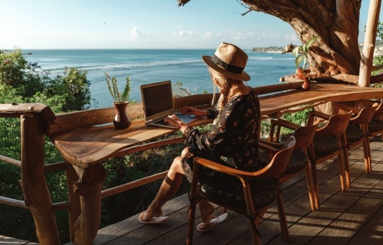Eine Arbeitnehmerin sitzt in einem Strandkaffee und arbeitet an ihrem Laptop.