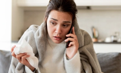 Eine erkältet wirkende Frau sitzt in eine Decke gehüllt auf einem Sofa, hält ein Taschentuch in der Hand und telefoniert.