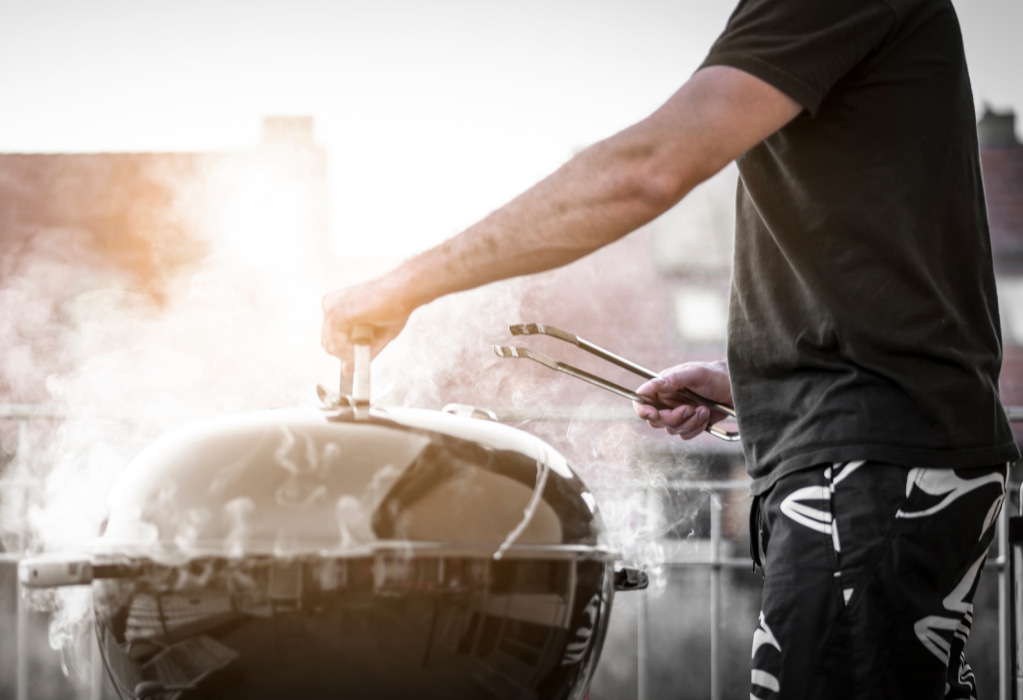 Ein Mann steht vor einem qualmenden Grill auf dem Balkon.
