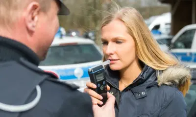 Eine Frau führt unter Aufsicht eines Polizisten einen Atemalkoholtest vor einem Fahrzeug durch.