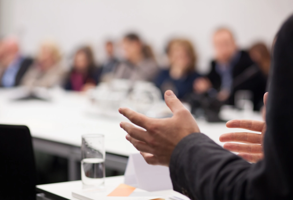 Der Fokus liegt auf den gestikulierenden Händen einer Person bei einer Rede oder Präsentation. Im Hintergrund sind mehrere Menschen zu sehen.