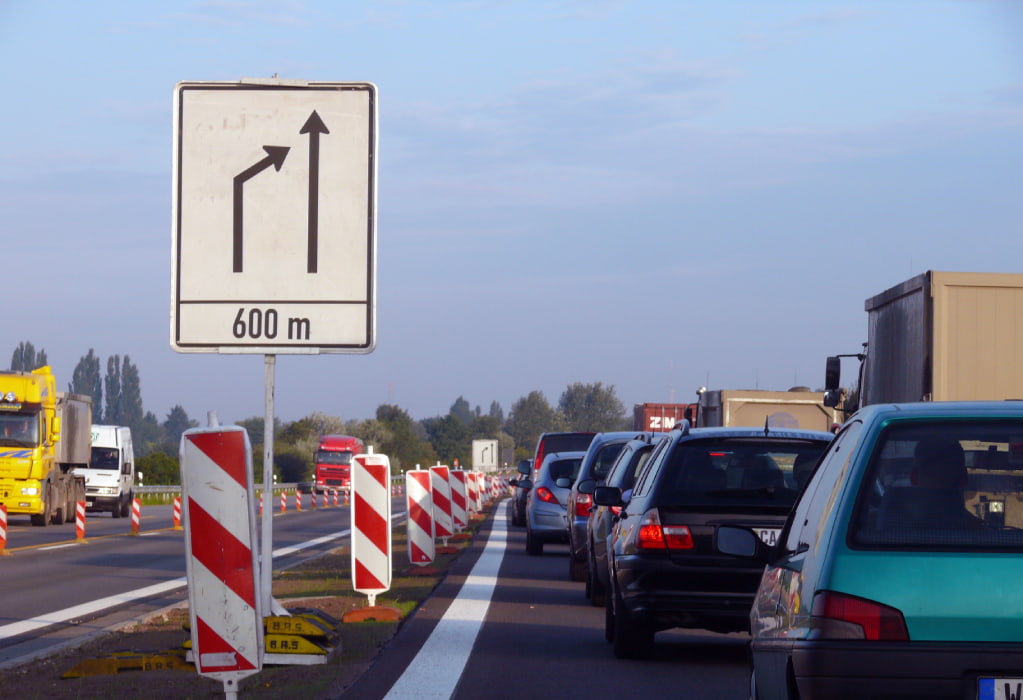 Ein Verkehrsschild kündigt das Ende der linken Fahrspur auf einer Autobahn an, was auf das Reißverschlussverfahren hinweist. Mehrere Fahrzeuge stehen im Stau.