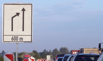 Ein Verkehrsschild kündigt das Ende der linken Fahrspur auf einer Autobahn an, was auf das Reißverschlussverfahren hinweist. Mehrere Fahrzeuge stehen im Stau.