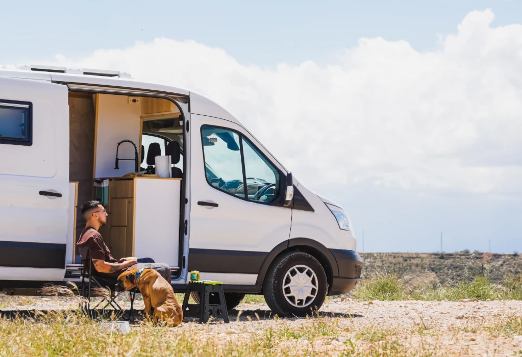 Ein Mann sitzt entspannt mit einem Hund vor einem Campervan in der Natur.