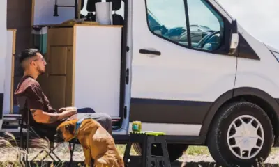 Ein Mann sitzt entspannt mit einem Hund vor einem Campervan in der Natur.