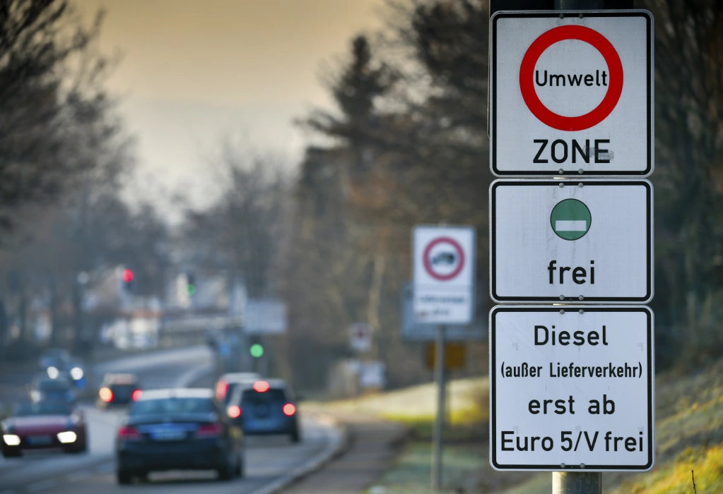Ein Straßenschild weist auf eine Umweltzone hin, in der nur Fahrzeuge mit grüner Plakette fahren dürfen. Im Hintergrund ist eine Straße mit mehreren Autos zu sehen. 