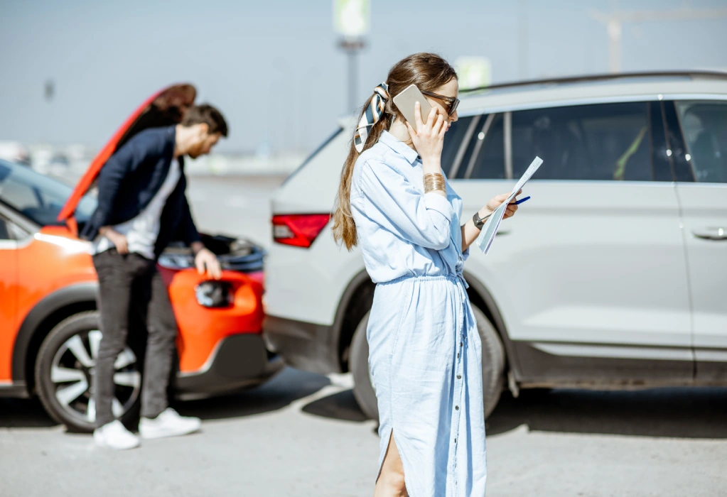 Eine Frau telefoniert und hält Unterlagen in der Hand, während im Hintergrund ein Mann den Schaden an zwei kollidierten Autos begutachtet.