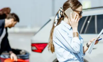 Eine Frau telefoniert und hält Unterlagen in der Hand, während im Hintergrund ein Mann den Schaden an zwei kollidierten Autos begutachtet.