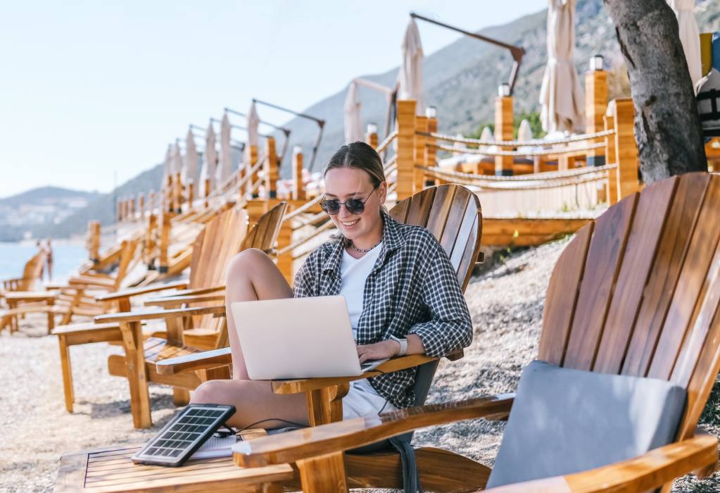 Eine Frau sitzt auf einem Stuhl am Strand und arbeitet entspannt mit einem Laptop.