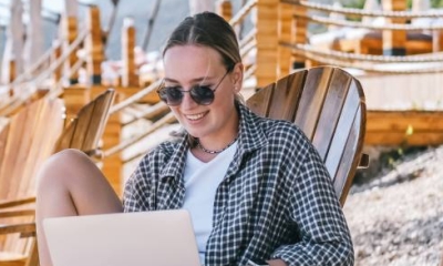 Eine Frau sitzt auf einem Stuhl am Strand und arbeitet entspannt mit einem Laptop.
